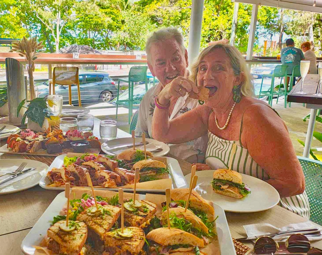 Two people enjoying sandwiches at an outdoor restaurant with greenery in the background.