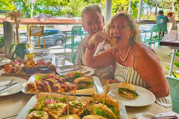 Two people enjoying sandwiches at an outdoor restaurant with greenery in the background.