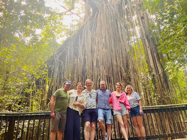 Group of six people standing in front of a large banyan tree in a forest.