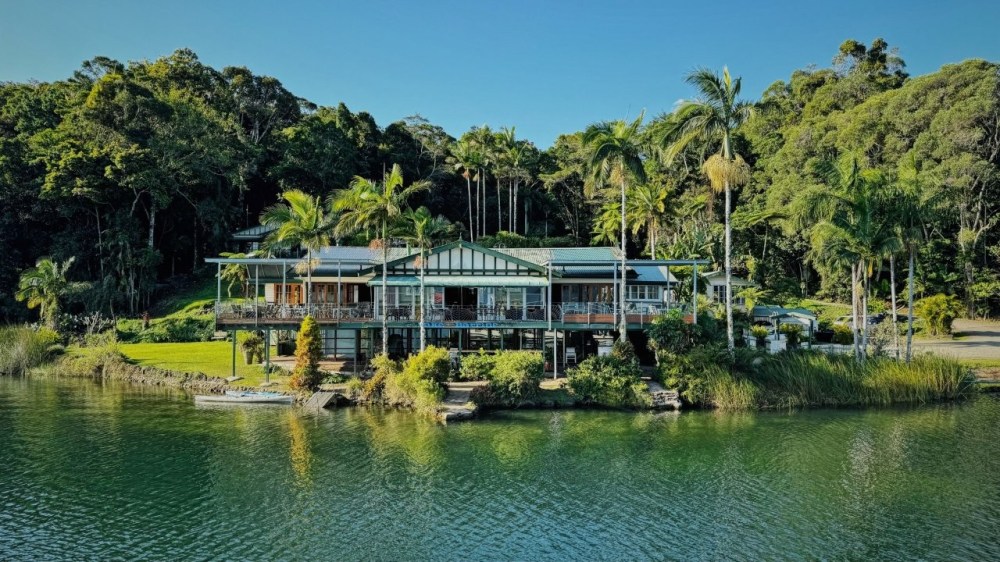 Large house surrounded by trees, located by a water body on a sunny day.
