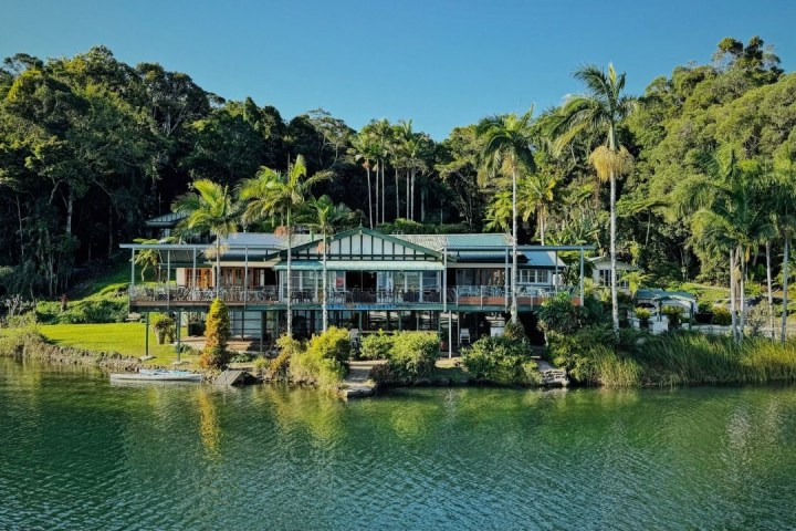 Large house surrounded by trees, located by a water body on a sunny day.