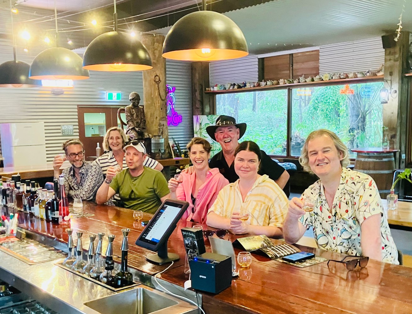 Group of people smiling at a bar counter, holding drinks; large pendant lights overhead.