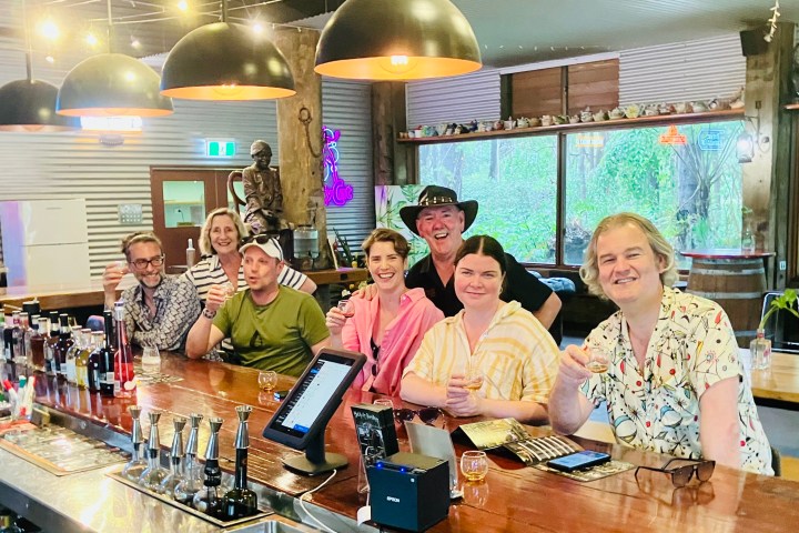 Group of people smiling at a bar counter, holding drinks; large pendant lights overhead.