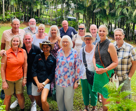Group of 14 smiling adults posing outdoors in front of palm trees.