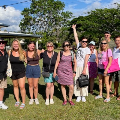 Group of people standing outdoors on grass, smiling, with trees and a house in background.