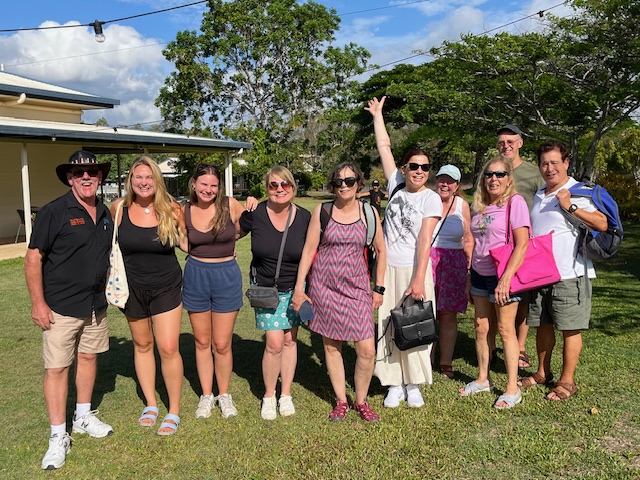 Group of people standing outdoors on grass, smiling, with trees and a house in background.