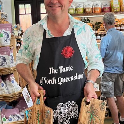 Man wearing an apron holds two decorated wooden boards in a store with products displayed around.