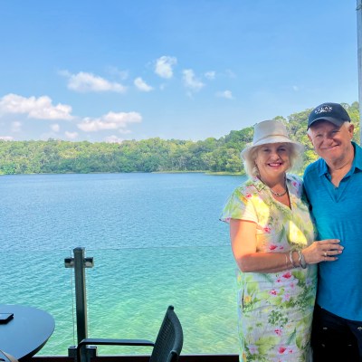 Smiling couple posing by a lake with forested shoreline under a clear blue sky.