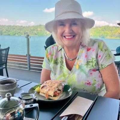 Woman in floral shirt and hat smiles at outdoor café table with sandwich and tea.