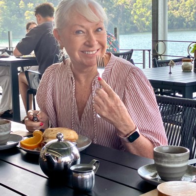 Smiling woman at table with tea, fruit, and pastry outdoors near water and forest.