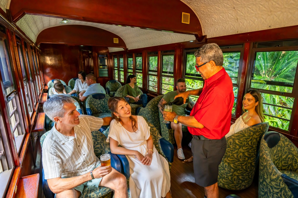 People in a train car enjoying drinks served by a man in a red shirt.