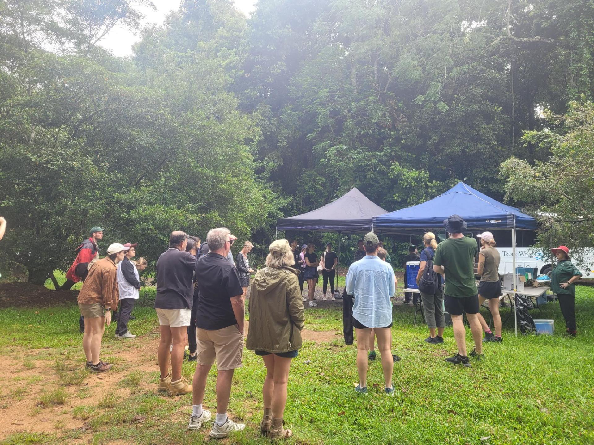 Group of people gathered outdoors under tents, surrounded by trees.