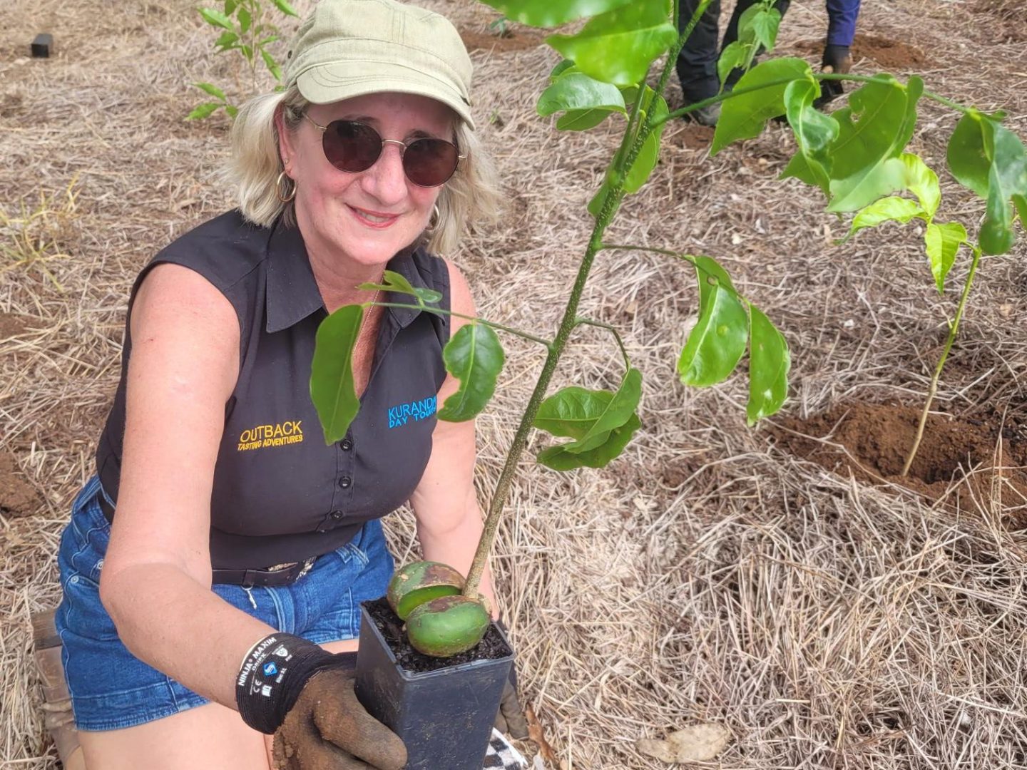 Woman kneeling with potted plant, wearing gloves and hat, in a group tree planting event.