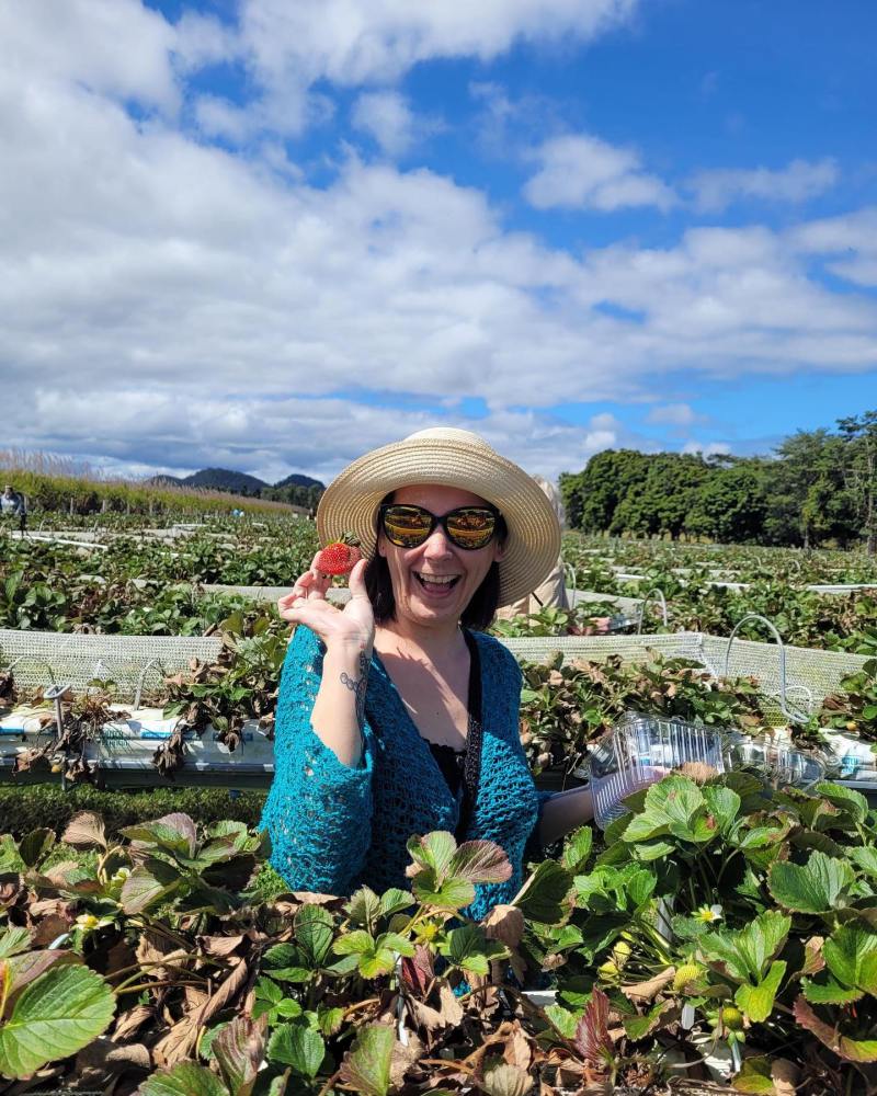 Person in a blue top and straw hat holds strawberry in a field under a partly cloudy sky.